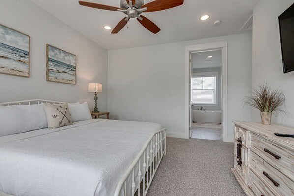 A bedroom with a bed, a ceiling fan, a nightstand with a lamp, a dresser, and a door leading to a bathroom.