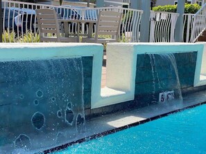 Una piscina con chorros de agua, sillas blancas y una valla al fondo.
