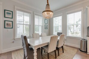 Une salle à manger avec une grande table, six chaises et un lustre. On y trouve trois fenêtres équipées de stores, et une poubelle est visible sur le côté.