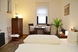 Quarto de hotel com cama, escrivaninha e duas cadeiras, radiador, planta em vaso e janela com persianas.