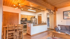 A modern kitchen with wooden cabinets, stainless steel appliances, and a central island. There is a dining area with wooden chairs and a sofa in the living room. - Easy Mountain - Mt Crested Butte (Crested Butte)