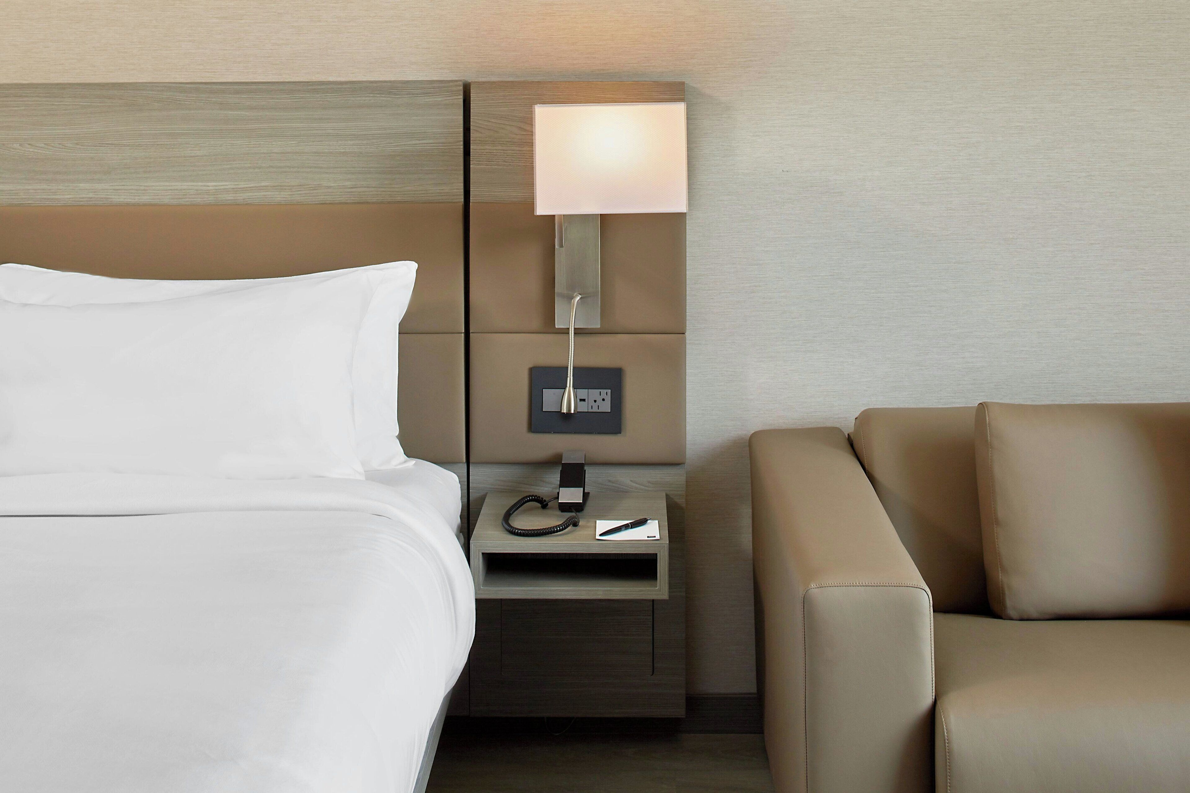 A hotel room with a bed, a nightstand with a lamp, a phone, and a remote control.