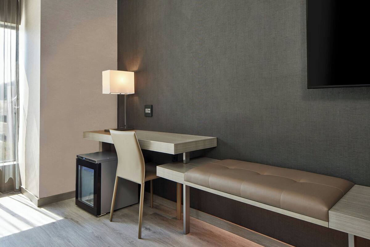 A modern hotel room with a desk, chair, bench, and a television mounted on the wall.