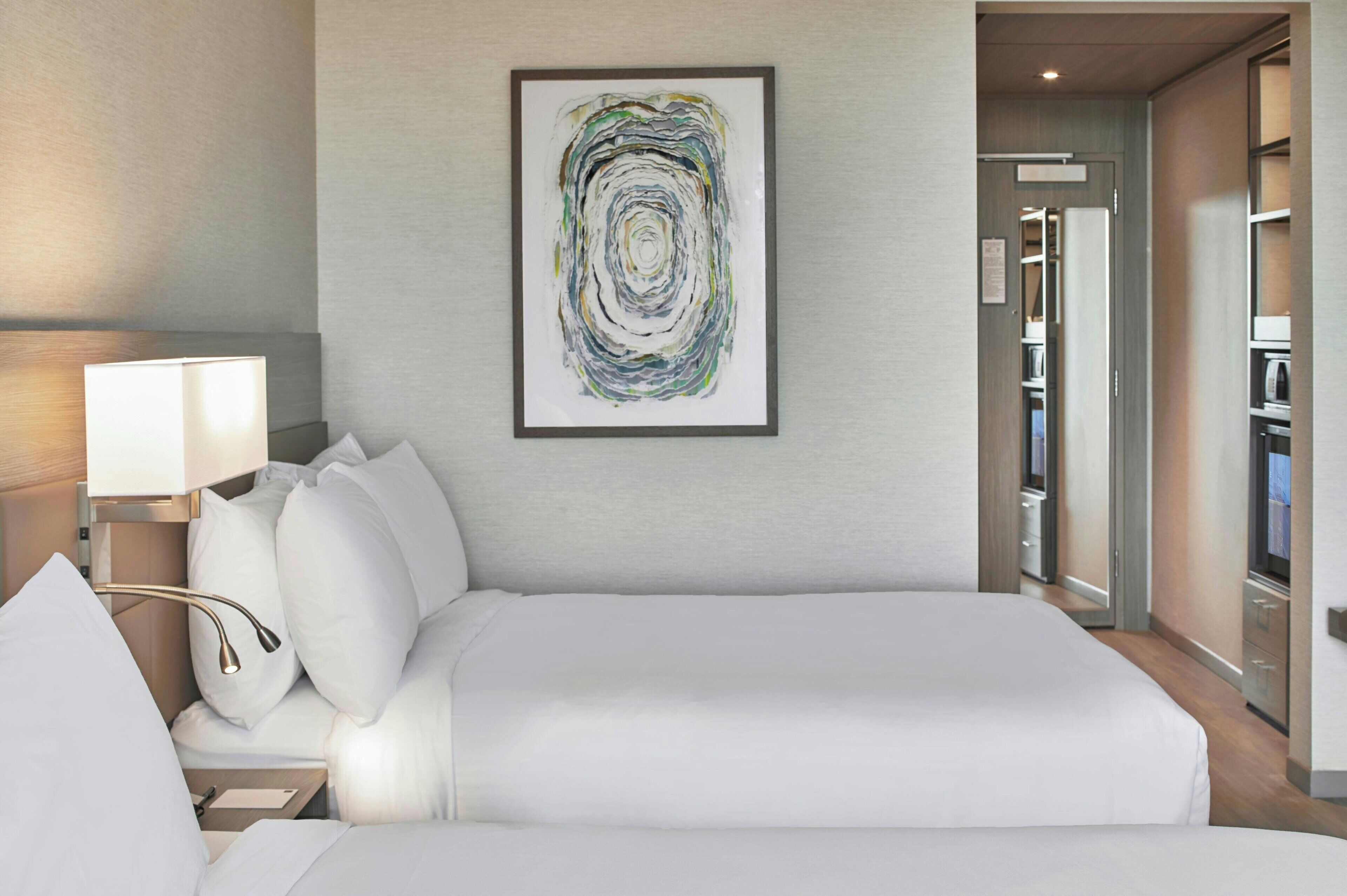 Un cuarto de hotel con una cama grande, una mesita de noche con lámpara, una obra de arte abstracta enmarcada en la pared y vistas al pasillo y al armario.