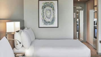 Un cuarto de hotel con una cama grande, una mesita de noche con lámpara, una obra de arte abstracta enmarcada en la pared y vistas al pasillo y al armario.