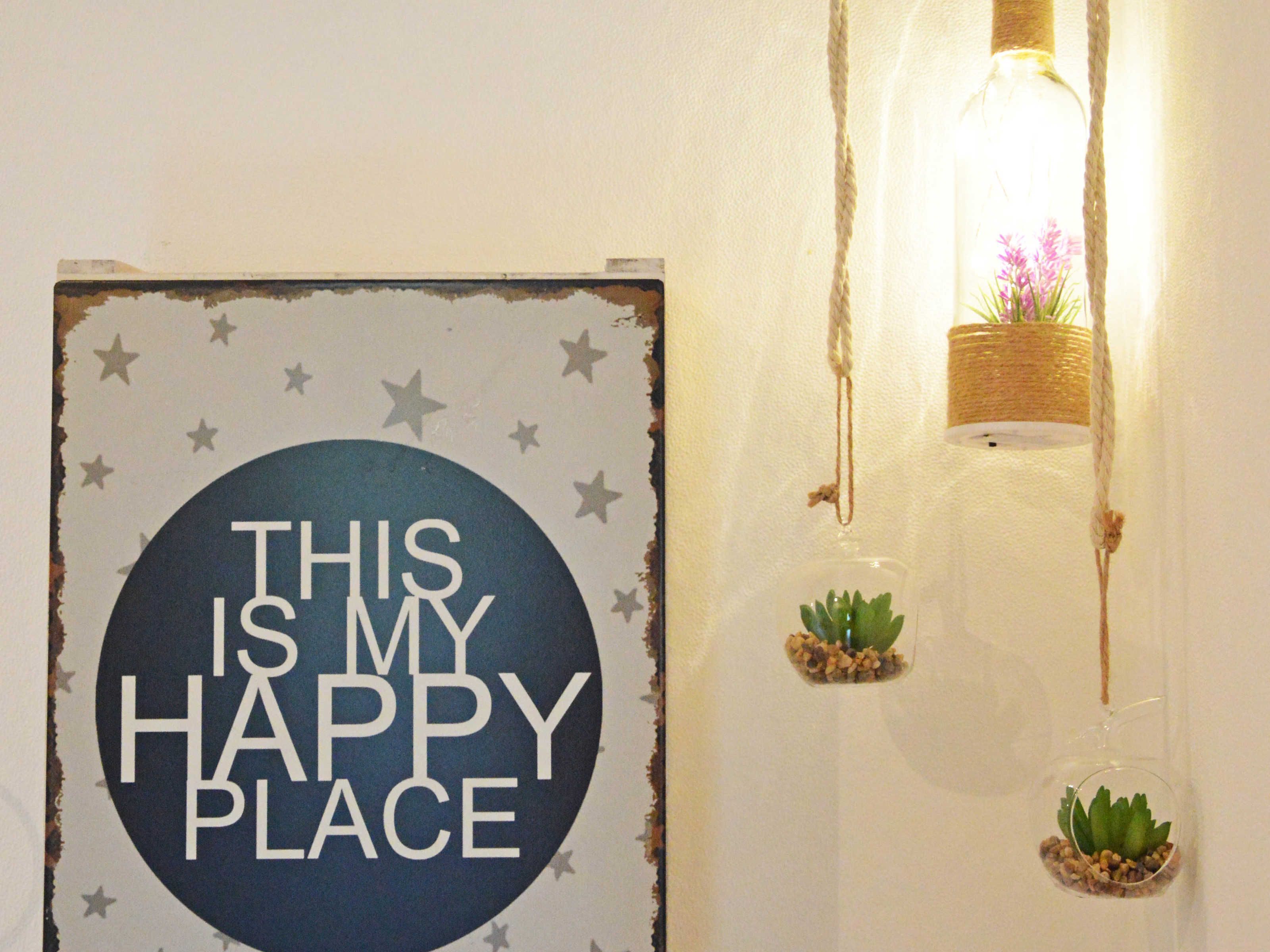 A wall with a circular sign that reads "THIS IS MY HAPPY PLACE" surrounded by stars. There are two hanging glass bottles with plants.
