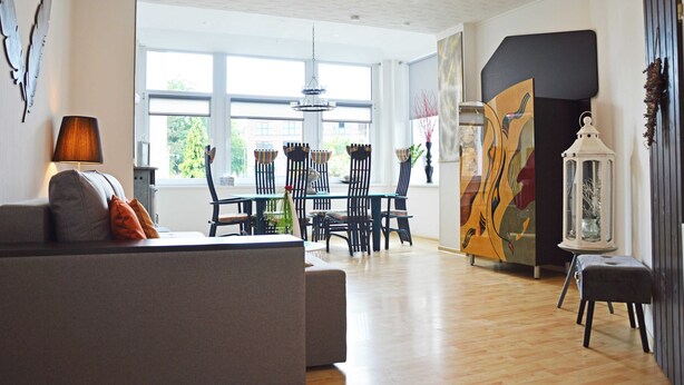 A modern living room with a sofa, dining table, and chairs. There is a large window providing natural light and a view of the outside. - Ferienwohnung Leuchtturm 29a - Ferienwohnung (Guben)