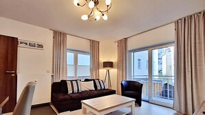 Una sala de estar moderna con un sofá, una mesa de centro, una lámpara de pie y un sillón. Hay un ventanal con cortinas, una puerta y un marco de cuadro en la pared.