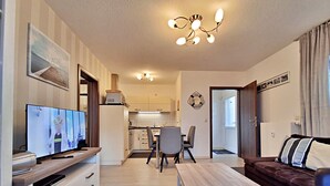 Una sala de estar moderna con televisor de pantalla plana, una mesa de centro de madera, un sofá de cuero marrón y un comedor con sillas, con una cocina al fondo.