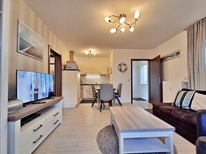 Un salon moderne avec un téléviseur à écran plat, une table basse en bois, un canapé en cuir marron et un coin repas avec des chaises, la cuisine étant visible au fond.