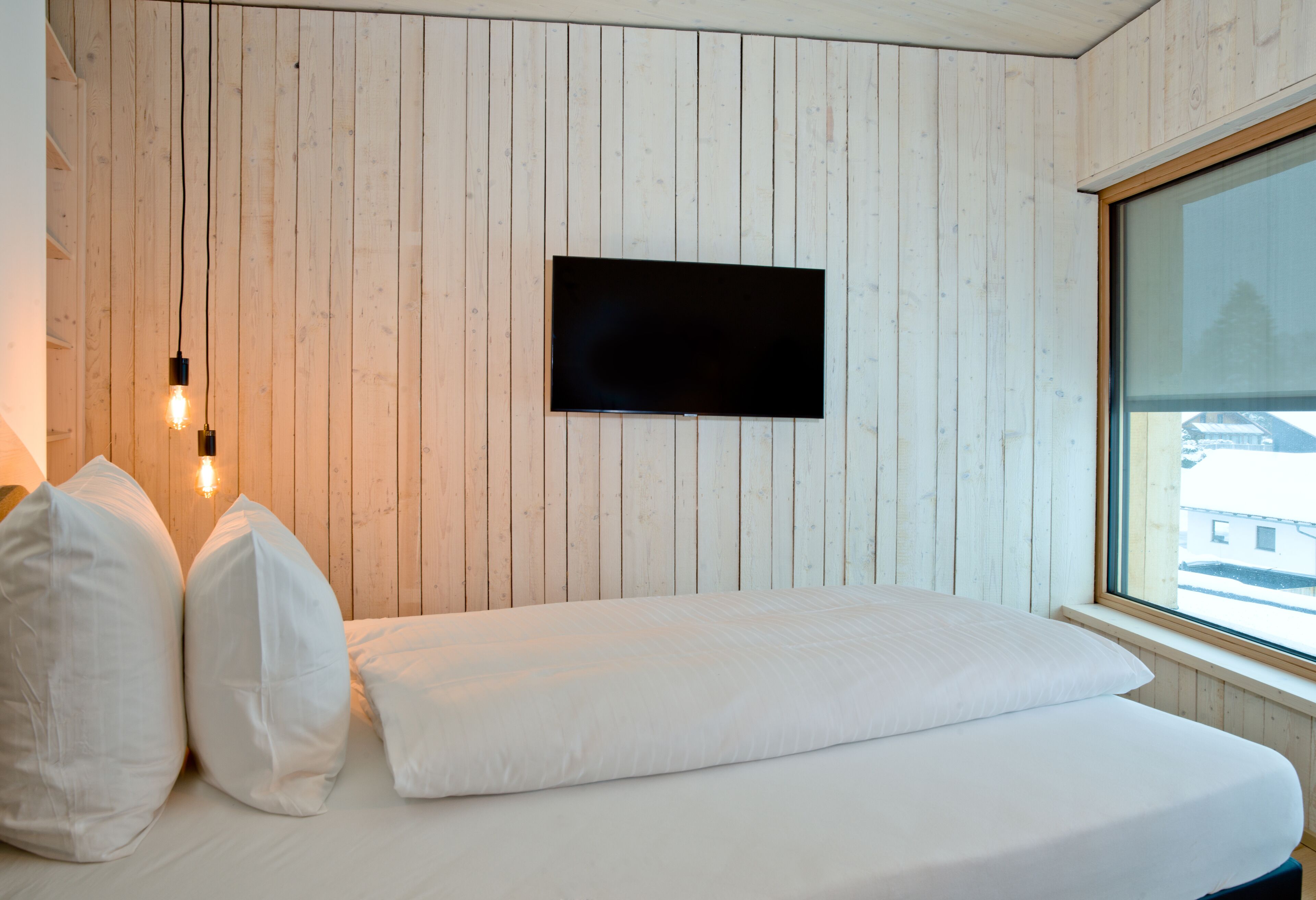 A bedroom with a wooden wall, a flat-screen TV mounted above a bed, and a window with a view of buildings outside.