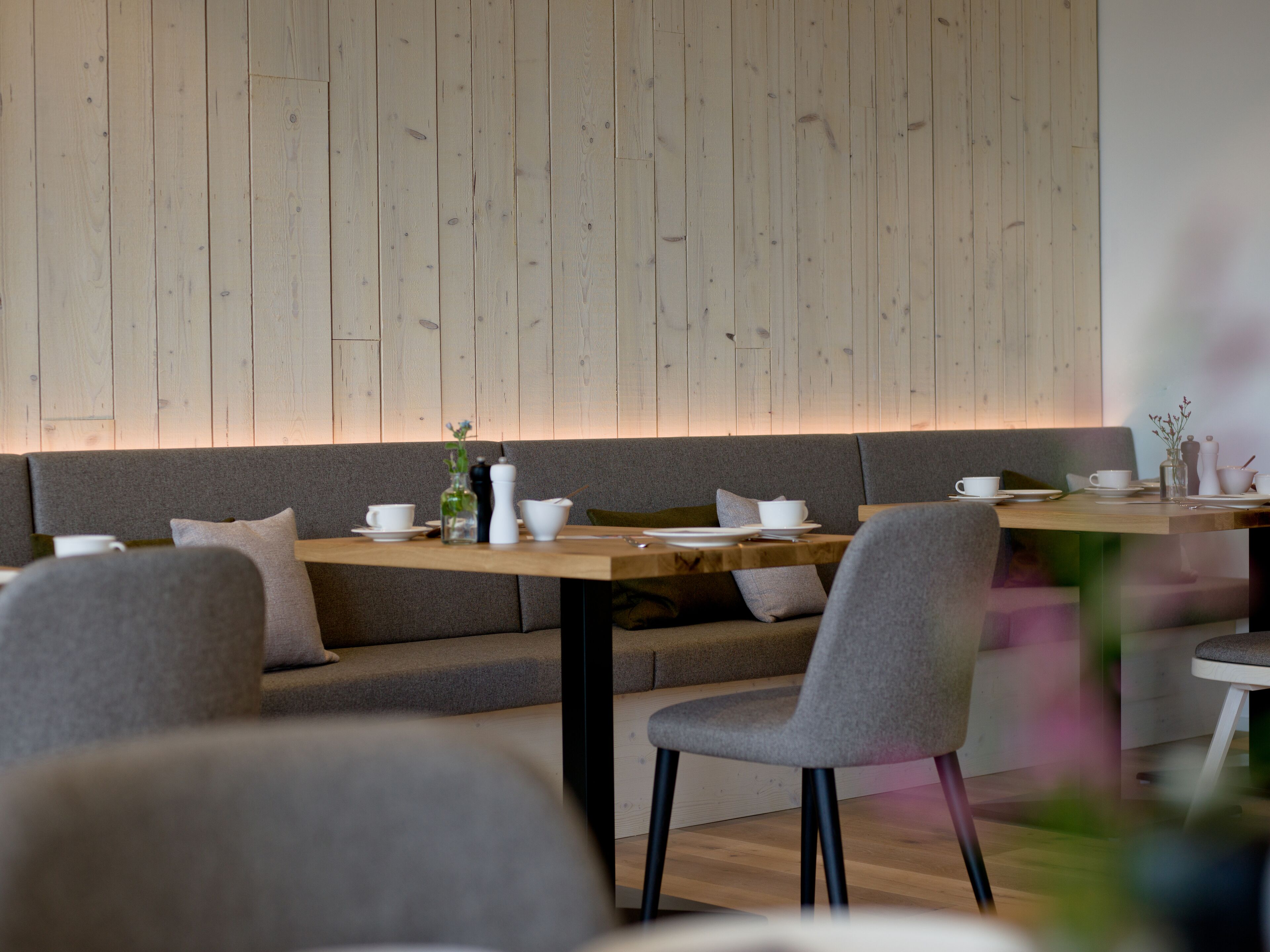 A modern dining area with a wooden wall, grey upholstered seating, and a wooden table set for two.