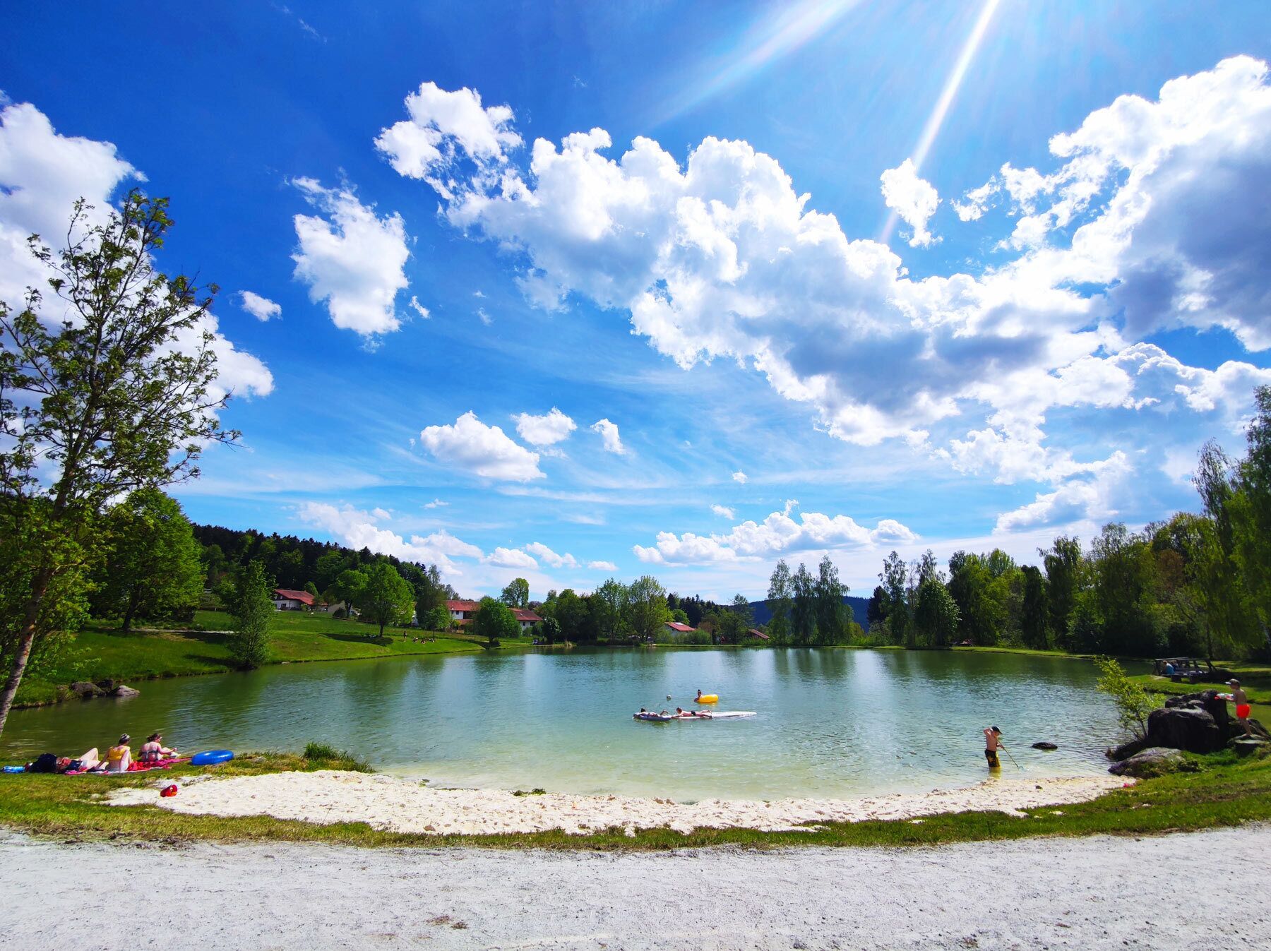 A lake with a sandy beach, surrounded by greenery and houses in the background.
