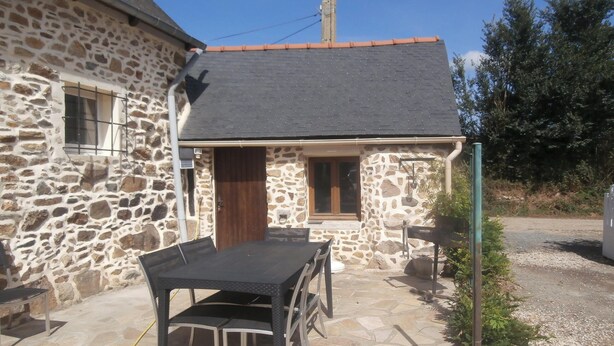 A stone building with a tiled roof, a wooden door, and a small window. There is a table and chairs outside.