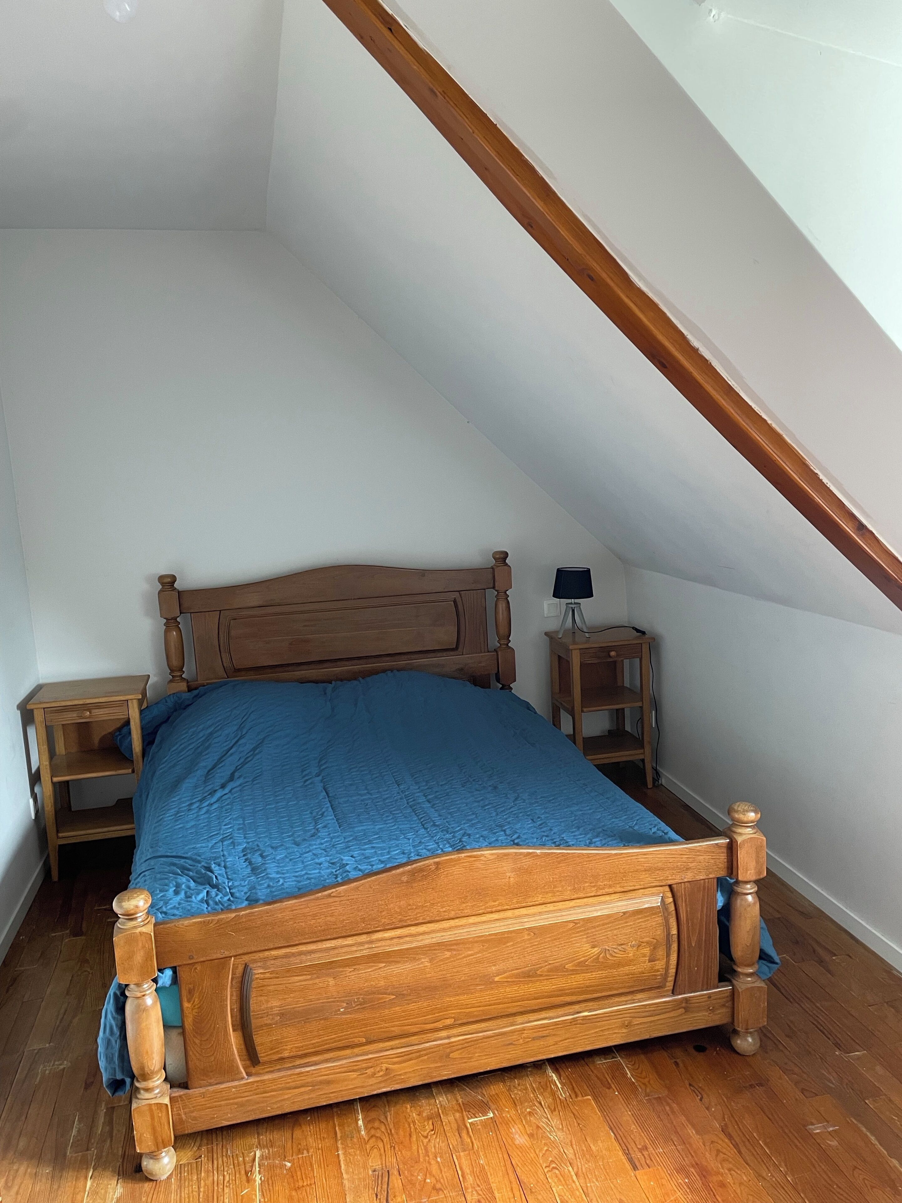 Ein Schlafzimmer mit schrägen Wänden, einem Bettgestell aus Holz, blauer Bettwäsche und einem Nachttisch mit Lampe.