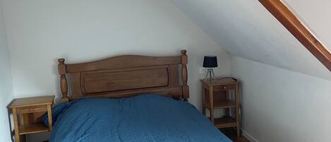 Ein Schlafzimmer mit schrägen Wänden, einem Bettgestell aus Holz, blauer Bettwäsche und einem Nachttisch mit Lampe.