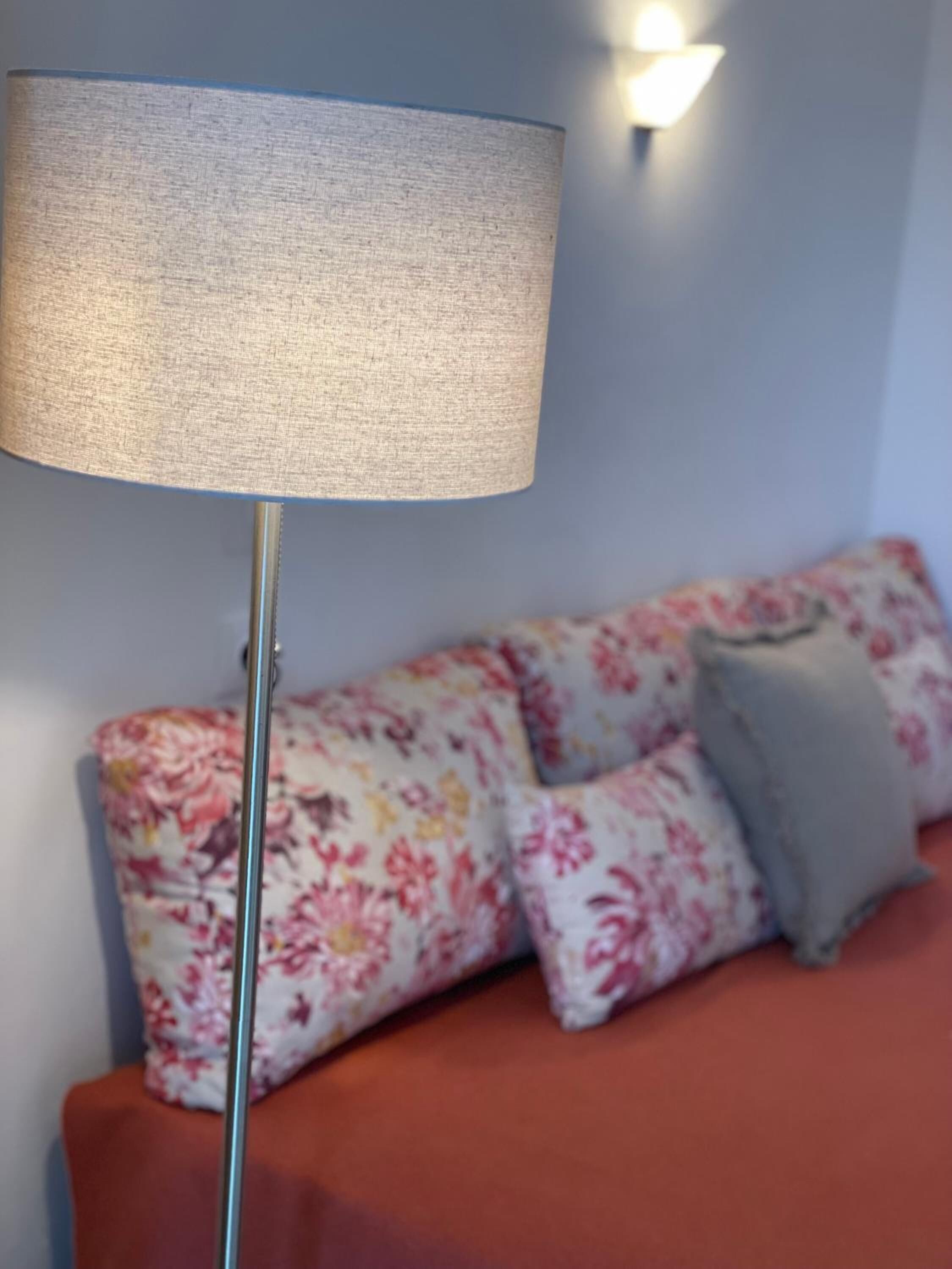 A room with a floor lamp, a sofa with floral cushions, and a wall-mounted light fixture.