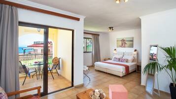 A hotel room with a balcony, a bed with a red blanket, a wooden table with a bowl of decorative items, and a potted plant.