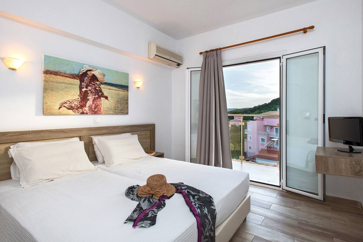 A hotel room with a large bed, a painting on the wall, a TV on a wooden stand, and a view of buildings through a glass door.