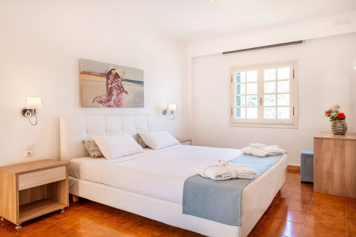 A bedroom with a large bed, a nightstand, a bedside table, a painting on the wall, and a window with a view.