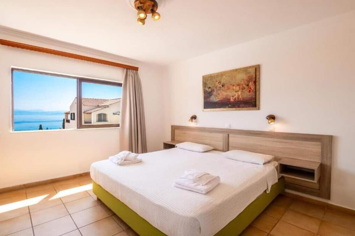 A hotel room with a large bed, a painting on the wall, and a view of the sea through a window.