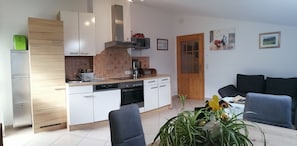 A modern kitchen with white cabinets, a built-in oven, and a coffee machine. There is a dining area with a black sofa and a table with a plant. - Panoramic View Vacation Apartment - Panoramic View Vacation Apartment (Götzens)