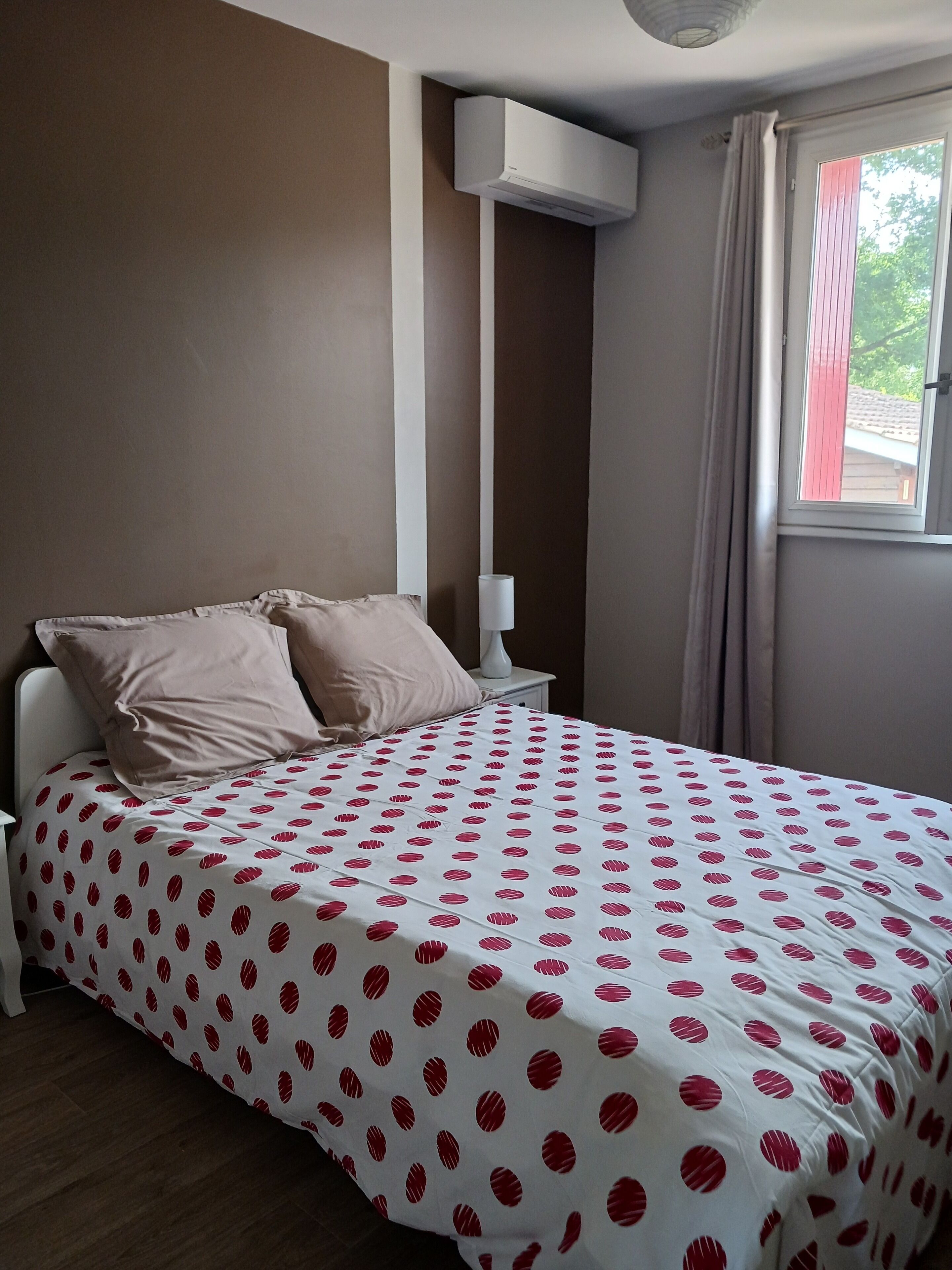 A bedroom with a bed, pillows, a nightstand, a lamp, an air conditioning unit, and a window with curtains.