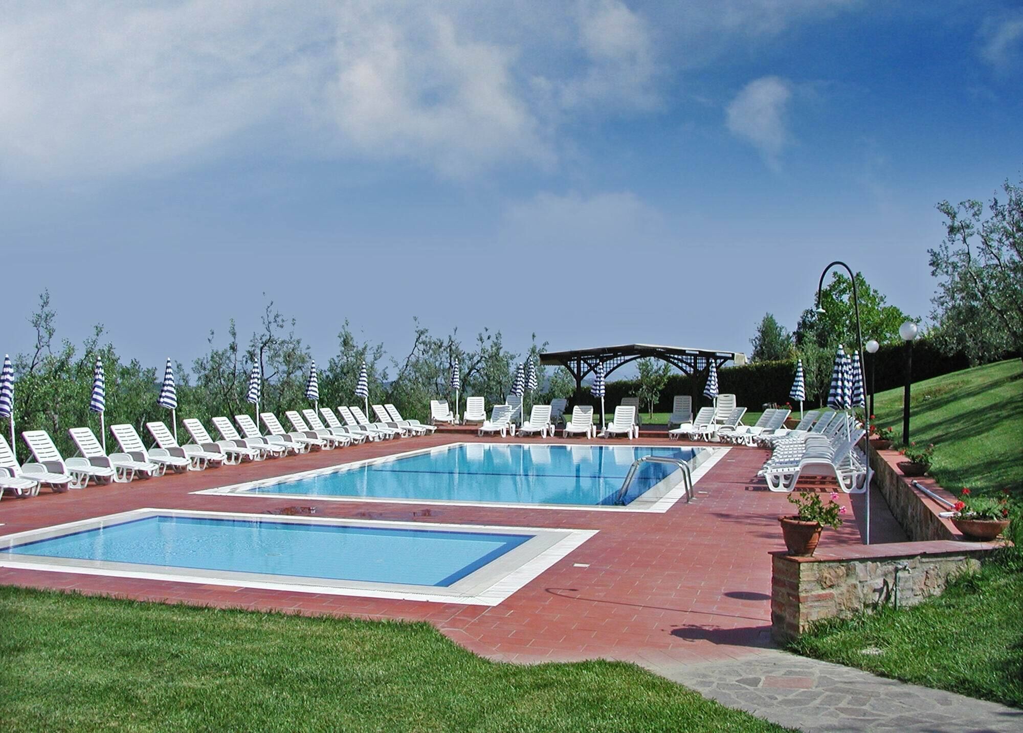 Un área de piscina con múltiples sillones de descanso y sombrillas, rodeada por un césped bien cuidado y una estructura de pérgola.