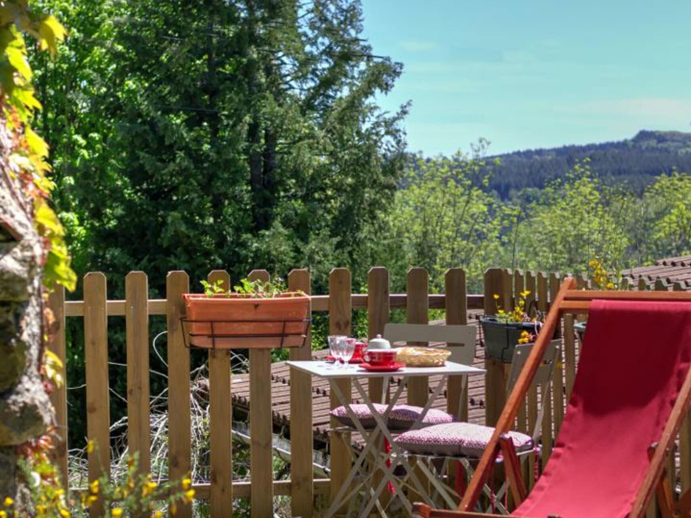 A wooden deck with a table and chairs, surrounded by a wooden fence and greenery.