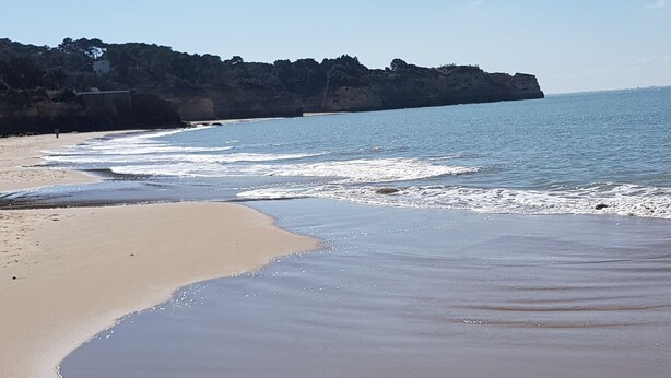 Una playa con una orilla curva, agua tranquila y un acantilado rocoso al fondo.