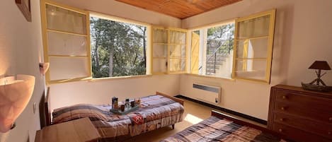 A cozy bedroom with a wooden ceiling, large windows with shutters, a bed with a patterned bedspread, a bedside table, and a dresser with a lamp.