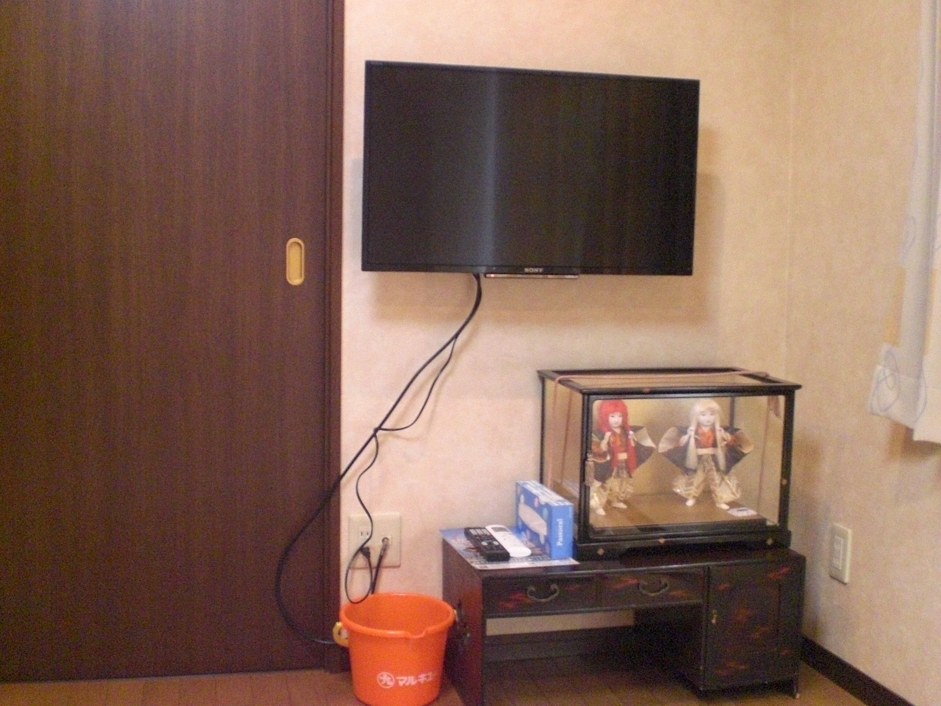 A television mounted on a wall, a small cabinet with a figurine, and an orange bucket.