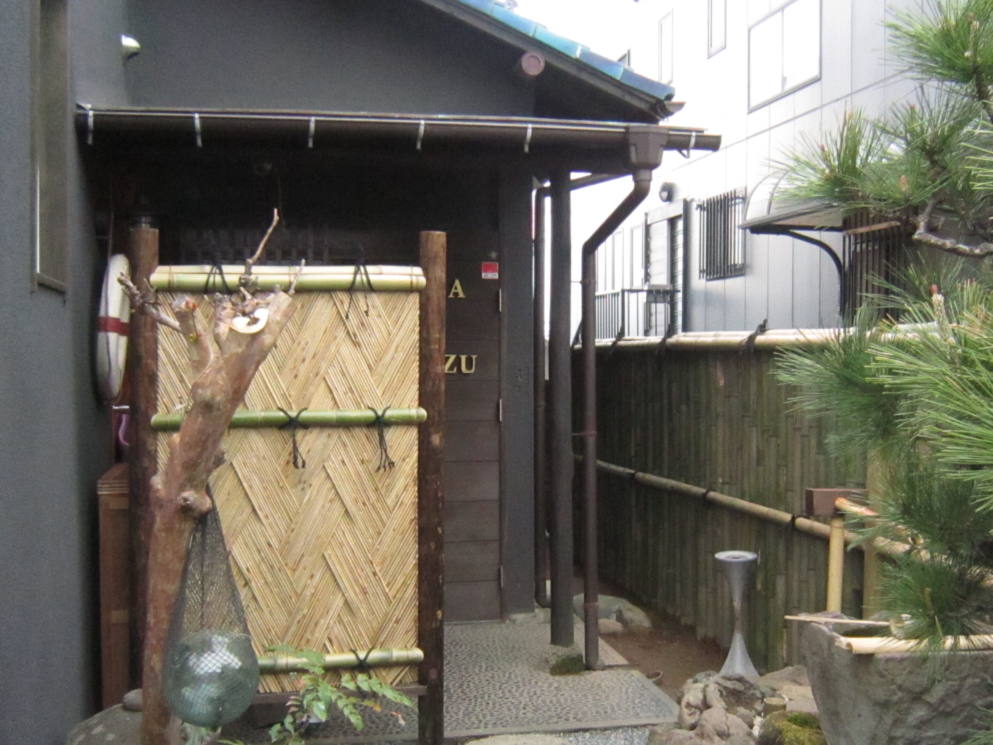 A wooden gate with a bamboo fence, a small tree, and a stone pathway leading to a building with a dark facade and a sign that reads "ZU".