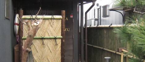 A wooden gate with a bamboo fence, a small tree, and a stone pathway leading to a building with a dark facade and a sign that reads "ZU".