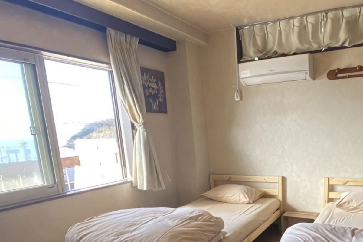 A room with two single beds, a window with curtains, a wall-mounted air conditioning unit, and a small table.