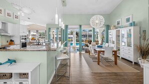 Eine moderne Küche mit weißen Schränken, ein Essbereich mit Holztisch und -stühlen sowie ein Blick auf einen Pool durch große Fenster.