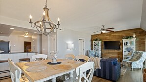 Eine moderne Küche mit einer Holzinsel, ein Essbereich mit Holztisch und weißen Stühlen, ein Wohnzimmer mit Sofa und Fernseher und ein Deckenventilator.