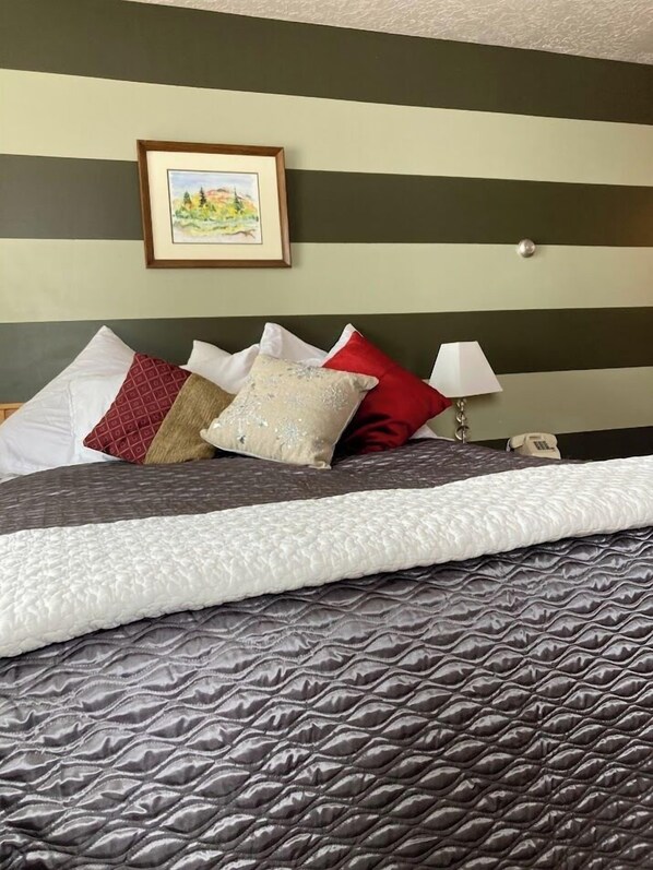 A neatly made bed with a patterned quilt, multiple pillows in various colors and patterns, a bedside lamp, and a framed picture on the wall. - Ski in/out studio room 105 at Powder Mountain's Columbine Inn. Sleeps 2 (Eden)