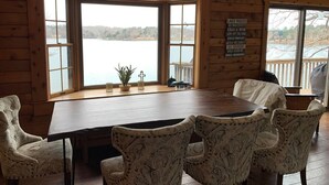 A wooden cabin with a large dining table and patterned chairs, large windows overlooking a lake, and a sign listing lake rules.