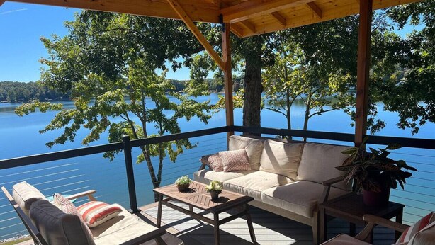 A covered outdoor seating area with a wooden deck, a sofa, and two chairs, overlooking a calm lake.