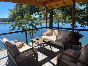Un espacio de estar al aire libre con techo, que cuenta con una terraza de madera, un sofá y dos sillas, con vista a un lago tranquilo.