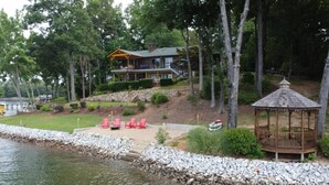 A lakeside property with a house, a gazebo, and red chairs by the water.