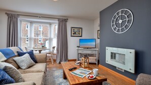 A living room with a sofa, coffee table, and a TV. There's a window with curtains, a clock on the wall, and a fireplace. - Oystercatcher (Whitby)