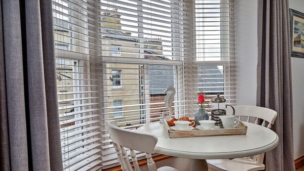 A dining area with a round table, white chairs, and a window with blinds. - Oystercatcher (Whitby)