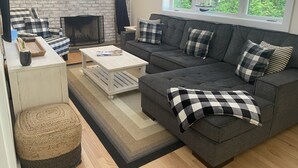 A living room with a fireplace, a sofa set, a coffee table, and a window with a view of greenery. - Brand New Charming Beach House (Mattituck)