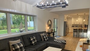 A modern living room with a grey sectional sofa, a white coffee table, and a kitchen area with bar stools. - Brand New Charming Beach House (Mattituck)