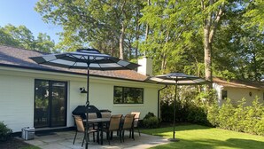A patio with a table and chairs, a grill, and two umbrellas. - Brand New Charming Beach House (Mattituck)