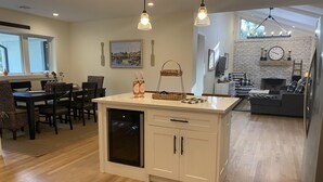 A modern kitchen with a central island, wooden flooring, pendant lighting, and a dining area with chairs and a table. - Brand New Charming Beach House (Mattituck)