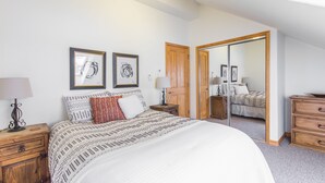 Un dormitorio con una cama grande, dos cuadros enmarcados, un armario de madera, una mesita de noche con lámpara y un espejo que refleja la habitación.