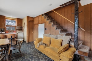 Ein gemütliches Blockhaus-Interieur mit Steinfußboden, Holzwänden, einer Couch mit Kissen, einem Esstisch mit Stühlen und einer Küchenzeile.